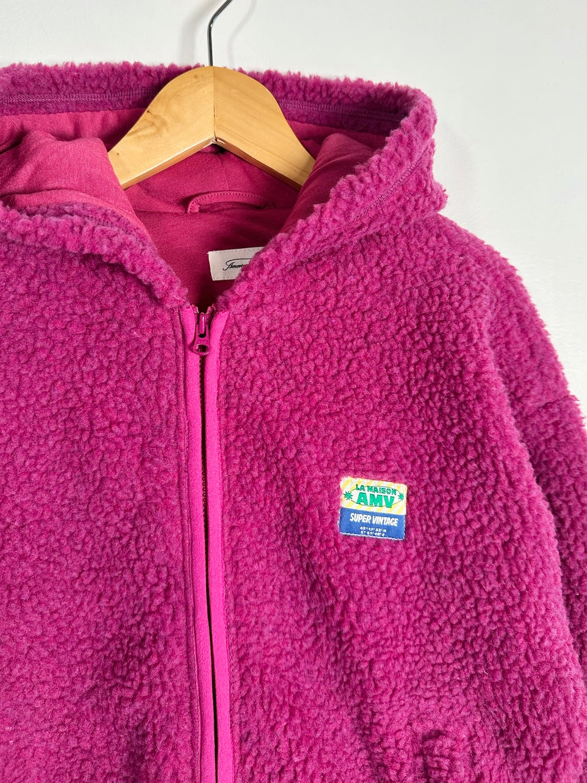A bright pink fleece zip-up hoodie with a hood and front zipper, hanging on a wooden hanger against a plain light background. A colorful 'La Maison Amy' patch is visible on the left chest.