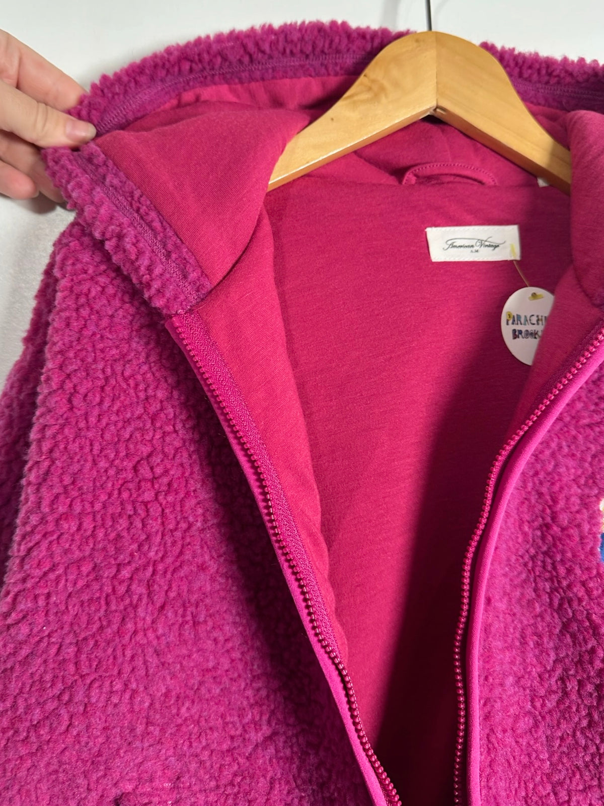 A close-up of a bright pink fleece zip-up jacket hanging on a wooden hanger, with a hand holding the collar open. The jacket has a textured sherpa/fleece exterior and a smoother inner lining, with a visible Tommy Hilfiger label.
