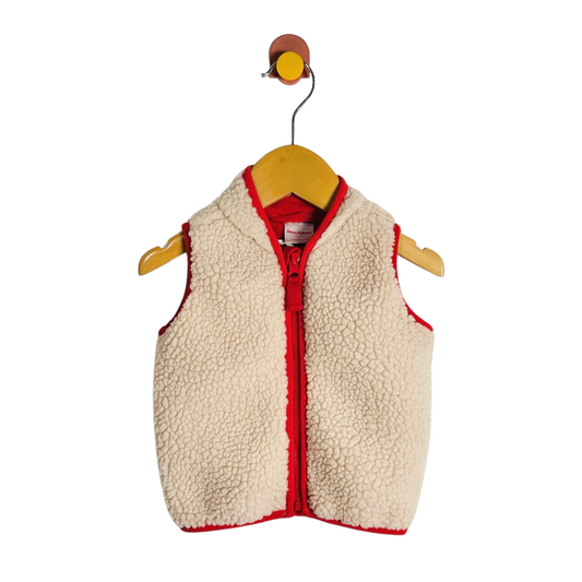 A cream sherpa fleece baby vest with red trim and a red zipper, hanging on a wooden hanger against a white background.