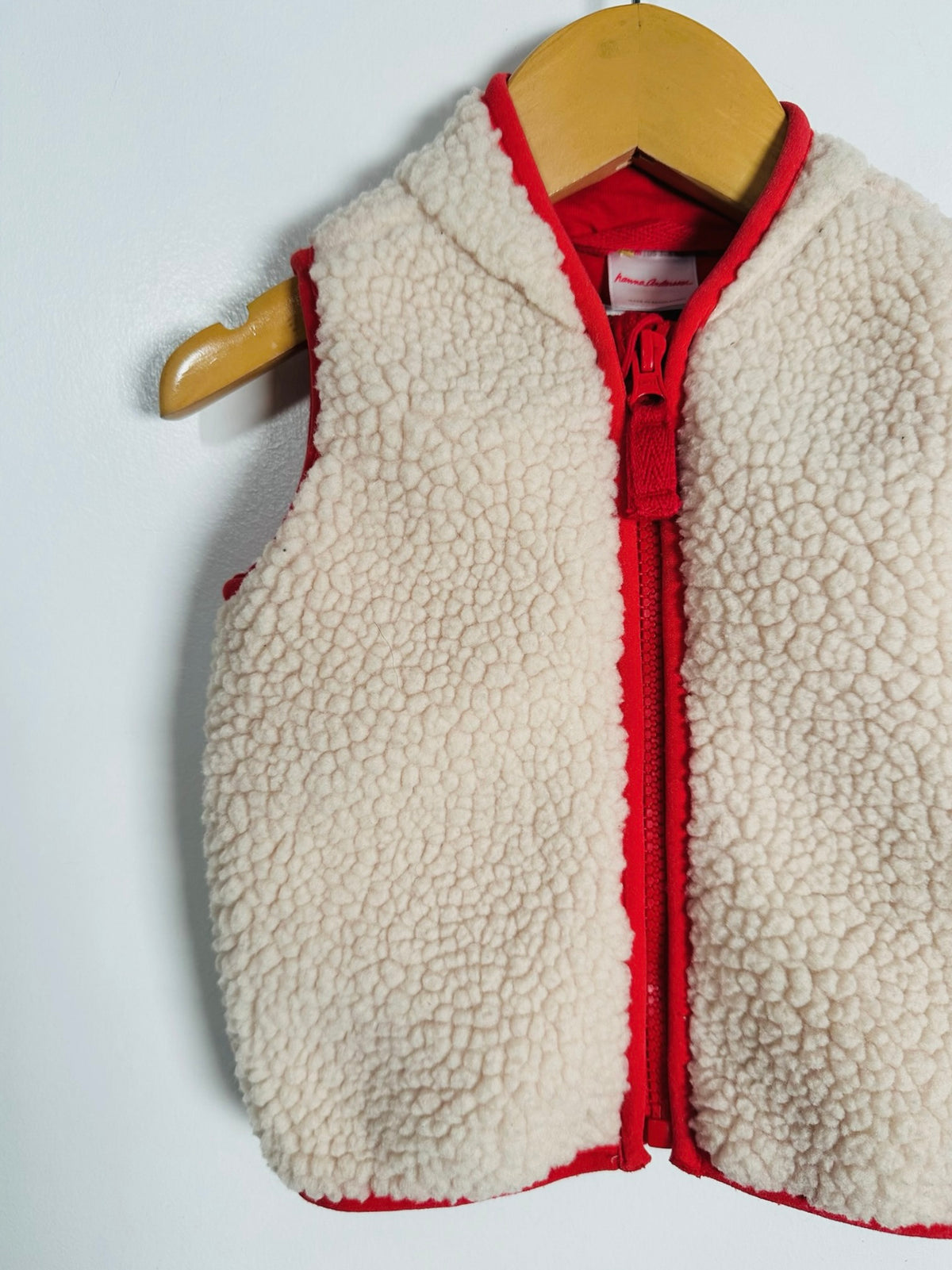 A cream sherpa fleece zip-up vest with red trim and a red zipper, hanging on a wooden hanger against a white wall.