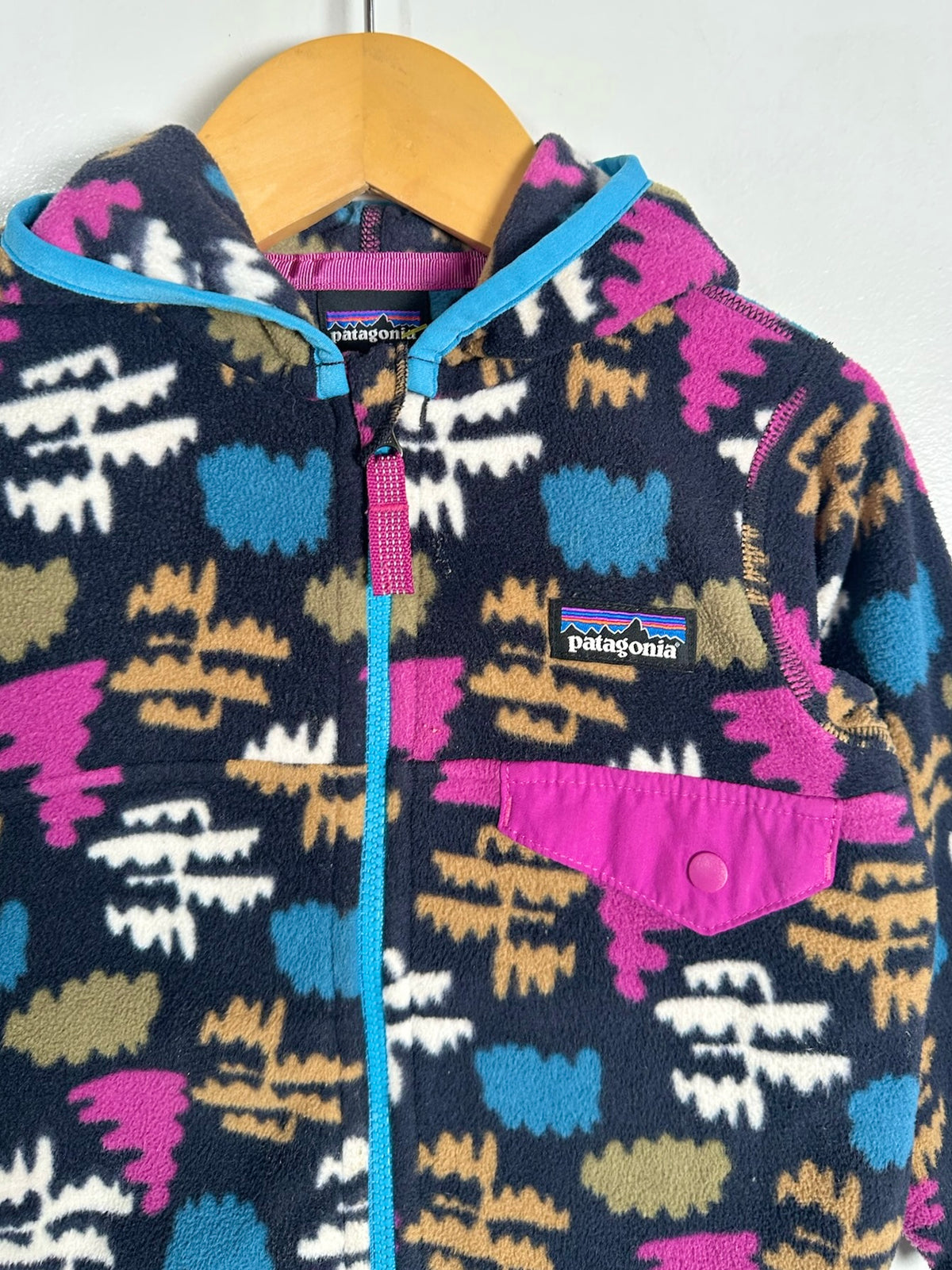 A close-up of a Patagonia fleece jacket hanging on a wooden hanger against a plain white wall. The jacket features a dark navy base with a colorful geometric mountain pattern in pink, blue, beige, and white, with a bright blue zipper and a pink chest pocket.