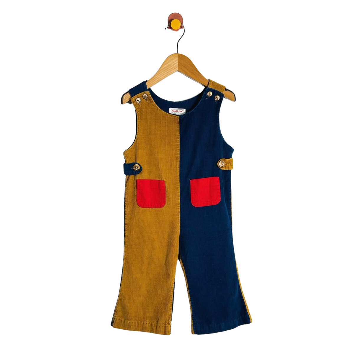 A children's sleeveless overall jumpsuit hanging on a wooden hanger against a white background. The garment is split vertically into two colors: mustard yellow corduroy on the left and navy blue on the right, with two contrasting red patch pockets and gold-tone metal buttons on the chest and waist.