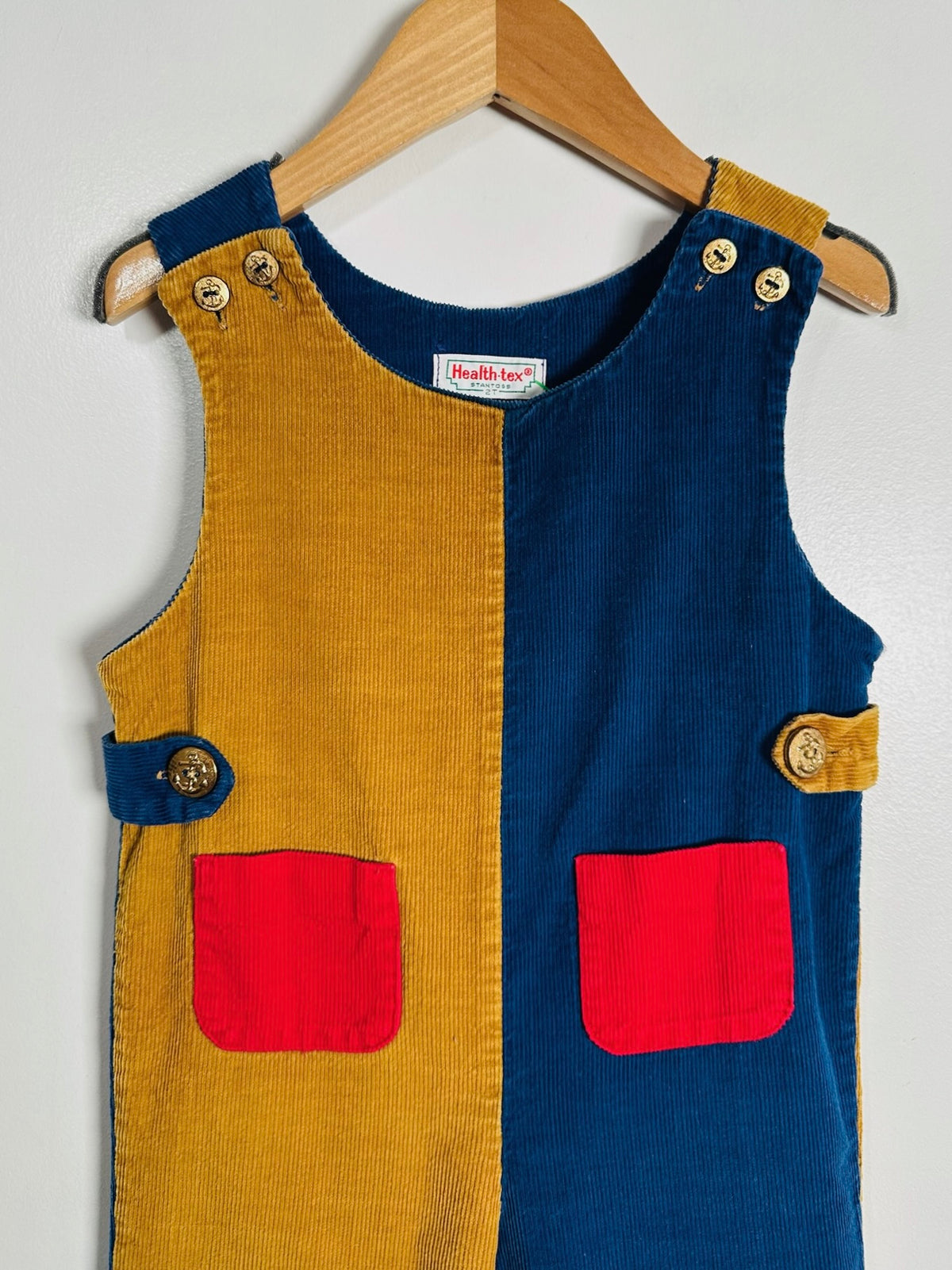 A children's sleeveless corduroy overall-style romper hanging on a wooden hanger against a plain light wall. The garment is split vertically into mustard yellow on the left and navy blue on the right, with red patch pockets and small decorative buttons.