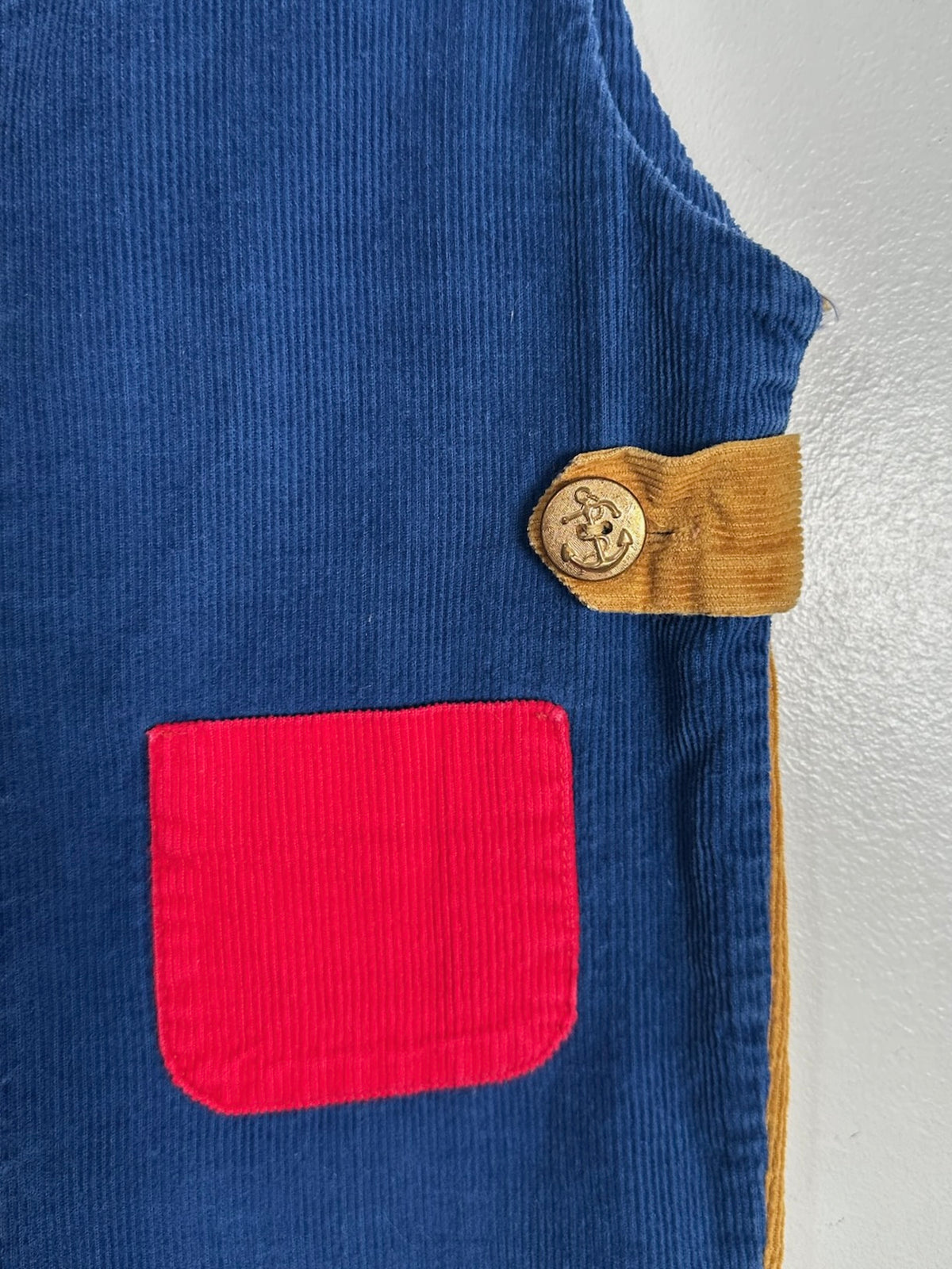 Close-up of a blue corduroy garment, likely a vest or jacket, featuring a bright red patch pocket and a tan corduroy tab with a circular metal emblem.