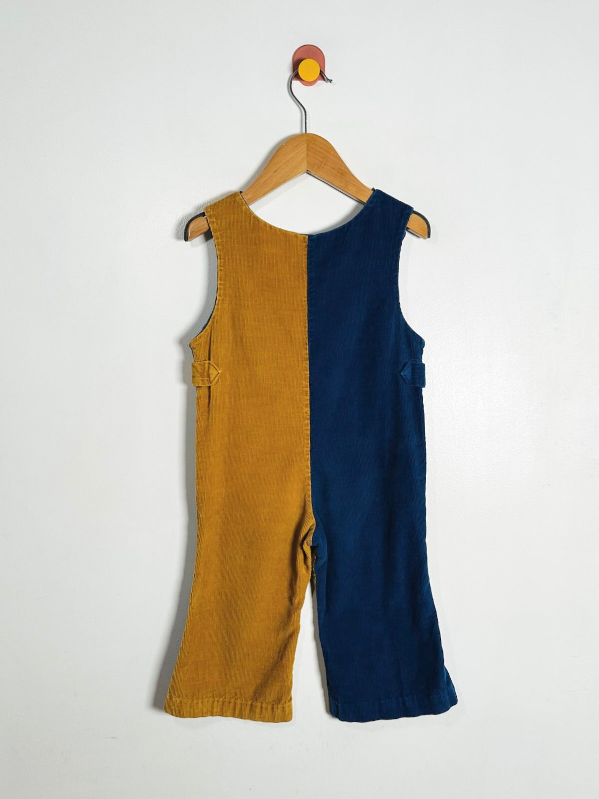 A sleeveless one-piece children's outfit hanging on a wooden hanger against a plain white wall. The garment is split vertically into two solid-color panels: mustard yellow on the left and navy blue on the right.