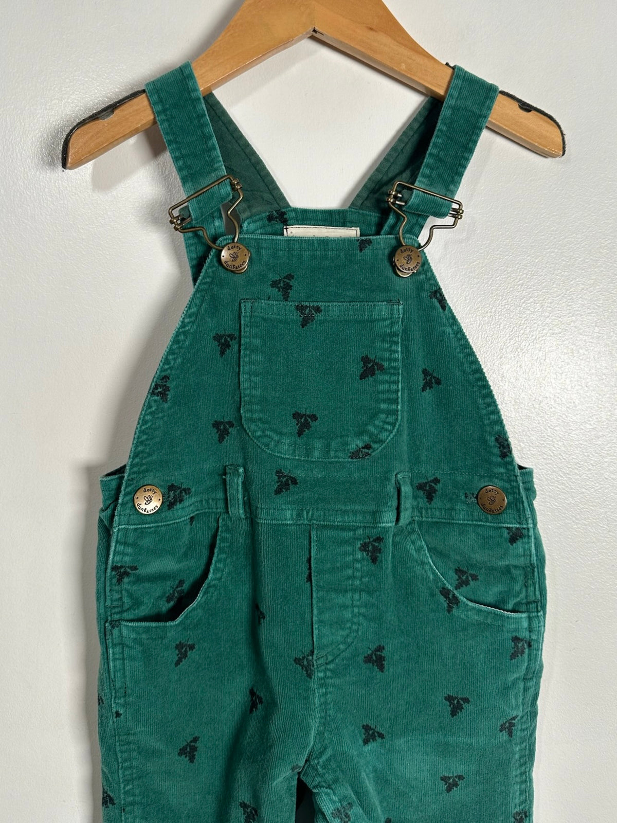 Green corduroy baby overalls with a black butterfly pattern, hanging on a wooden hanger against a white wall.