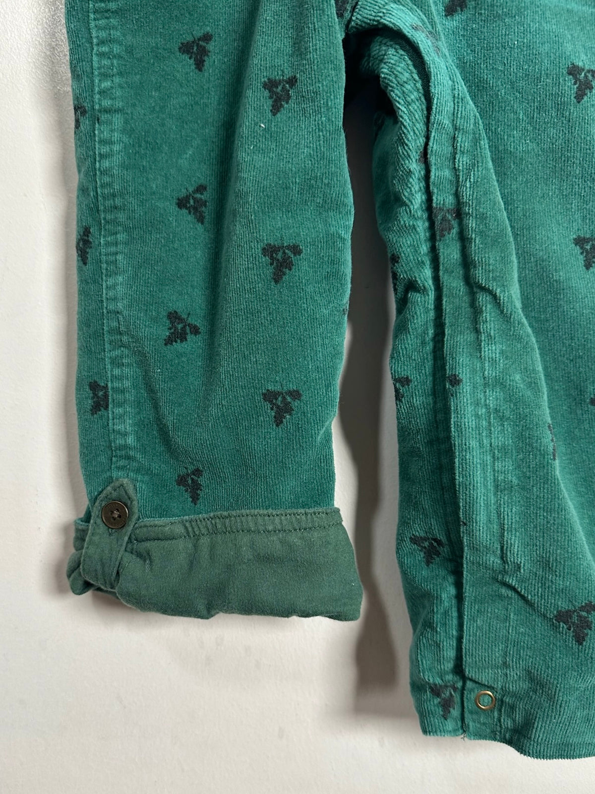 Close-up of a green corduroy garment, likely a jacket or overshirt, showing the sleeve cuff with a button and a repeating dark embroidered motif on the fabric.