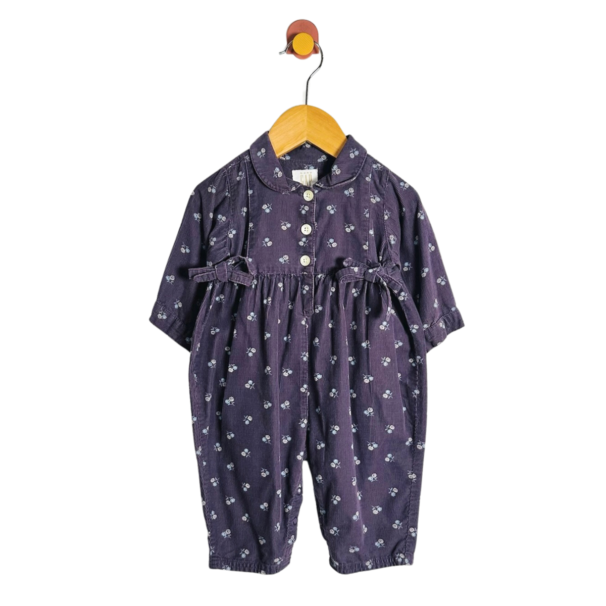 A baby girl's short-sleeve jumpsuit in dark purple with a small white floral print, hanging on a wooden hanger against a plain white background.