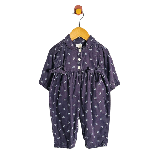 A baby girl's short-sleeve jumpsuit in dark purple with a small white floral print, hanging on a wooden hanger against a plain white background.