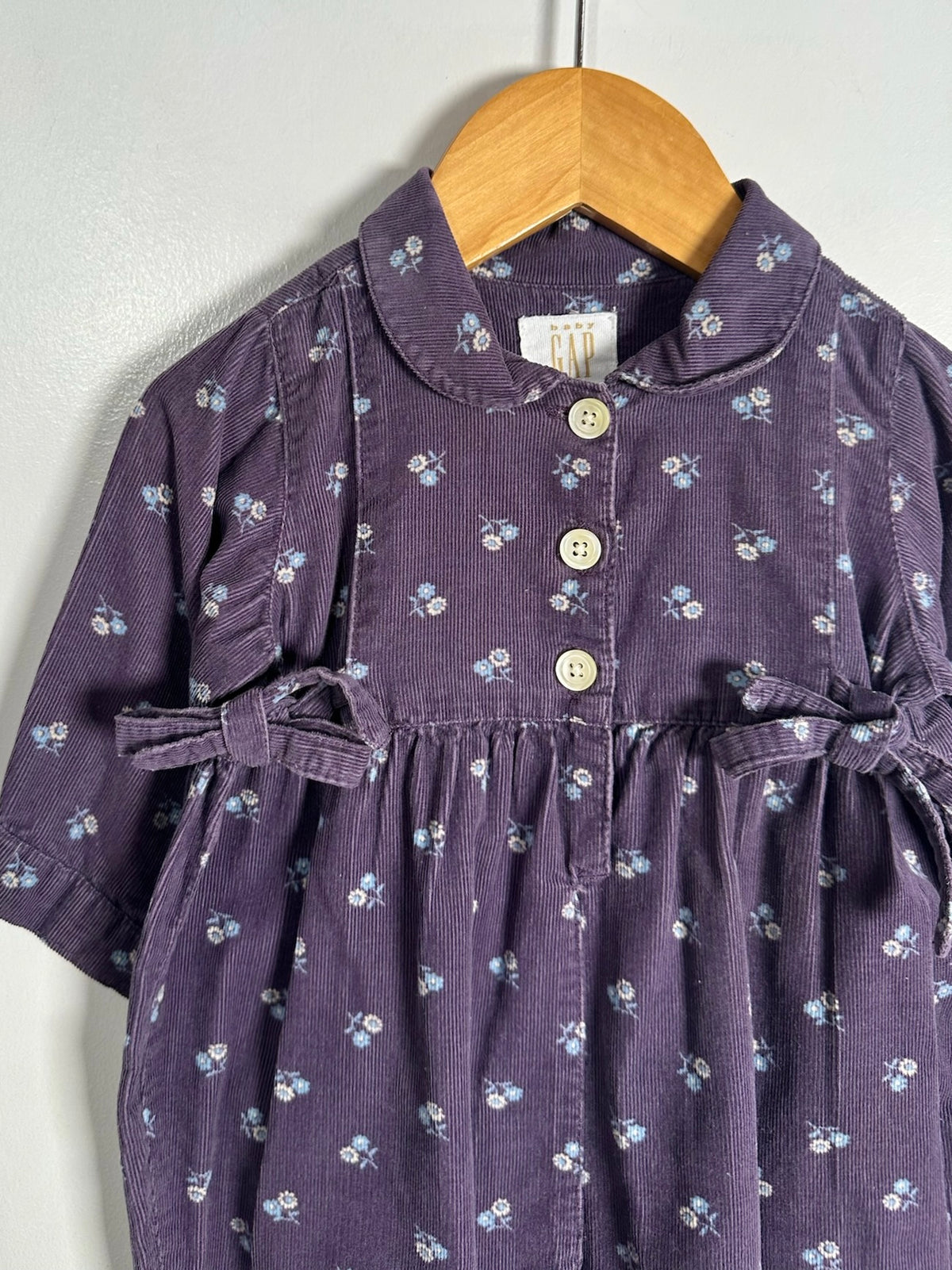 A toddler girl's short-sleeve corduroy dress in dark purple with a small white floral print, featuring a Peter Pan collar, three front buttons, and side bows, hanging on a wooden hanger against a light gray wall.