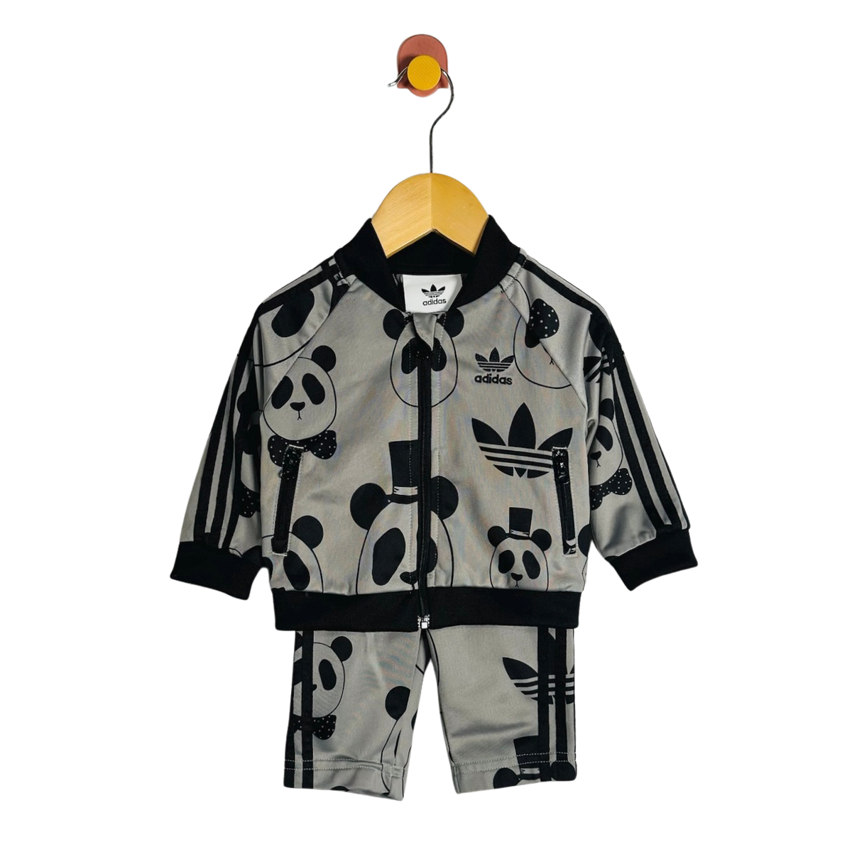 A children's two-piece tracksuit by adidas, featuring a light gray zip-up jacket and matching shorts with a black panda and trefoil logo print, hanging on a wooden hanger against a white background.