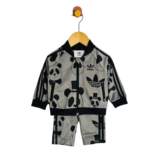 A children's two-piece tracksuit by adidas, featuring a light gray zip-up jacket and matching shorts with a black panda and trefoil logo print, hanging on a wooden hanger against a white background.