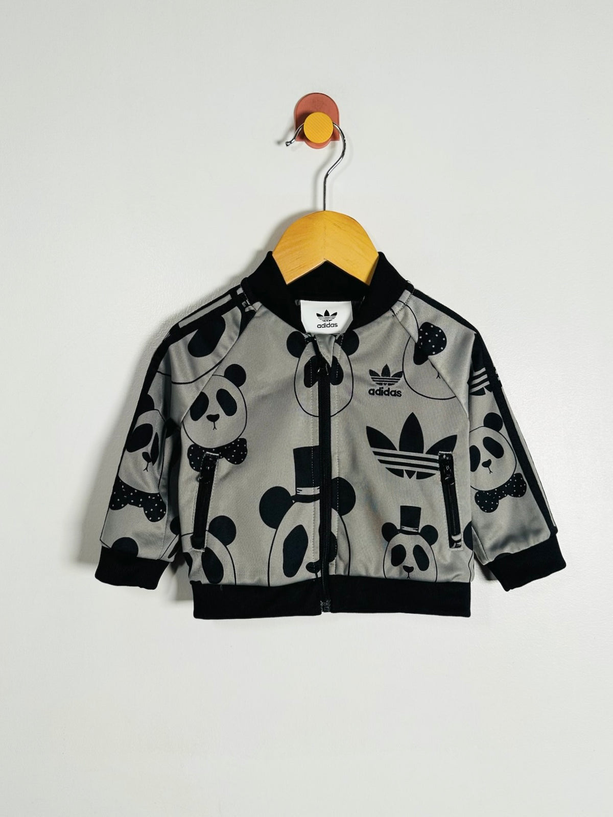 A children's gray bomber jacket with a black panda print, hanging on a wooden hanger against a plain white wall. The jacket features black ribbed collar, cuffs, and hem, with the Adidas trefoil logo and brand name visible on the chest.