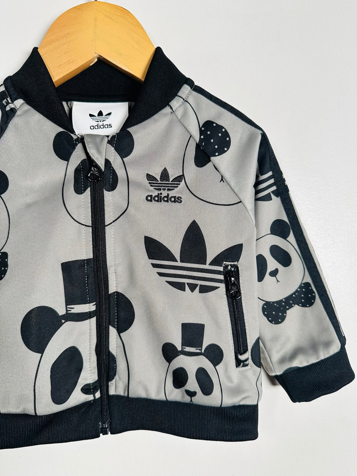 A children's Adidas zip-up jacket in light gray with a black panda and logo print, hanging on a wooden hanger against a plain light background.