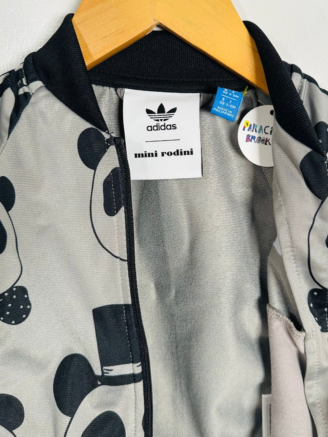 A close-up of an Adidas mini rodini jacket featuring a black and white Mickey Mouse pattern on a light gray background, hanging on a wooden hanger.
