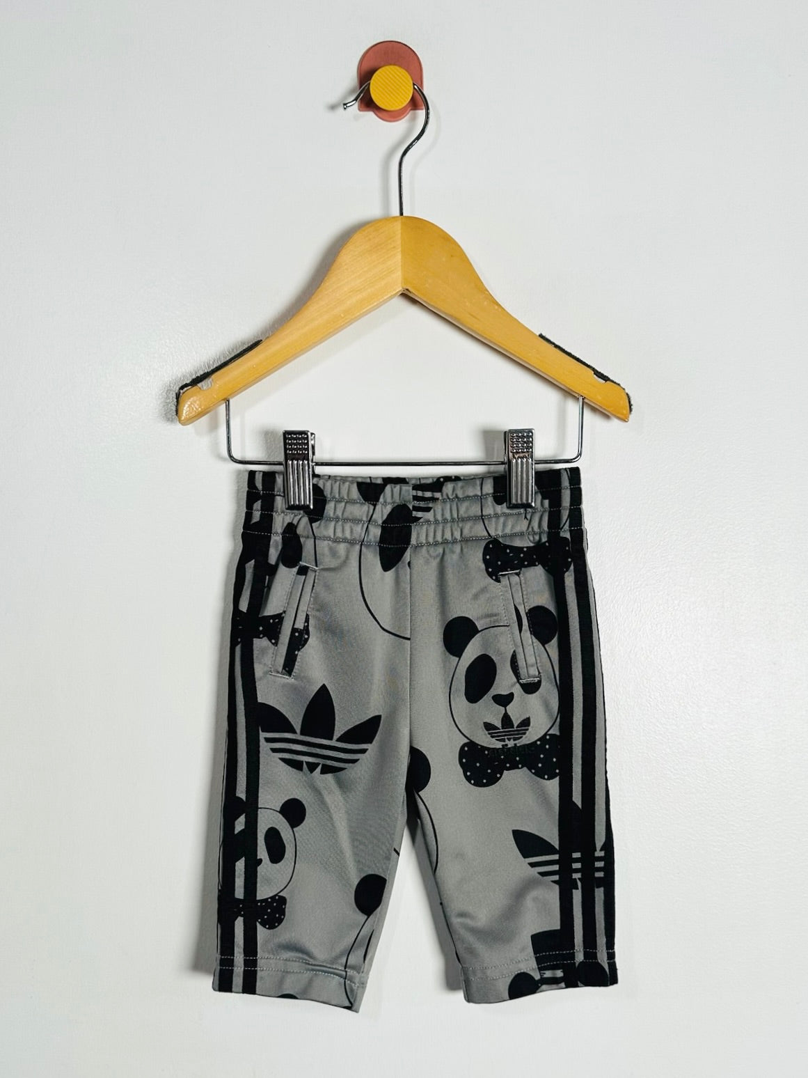 Gray Adidas track pants with a black panda print and signature three stripes, hanging on a wooden hanger against a plain white wall.