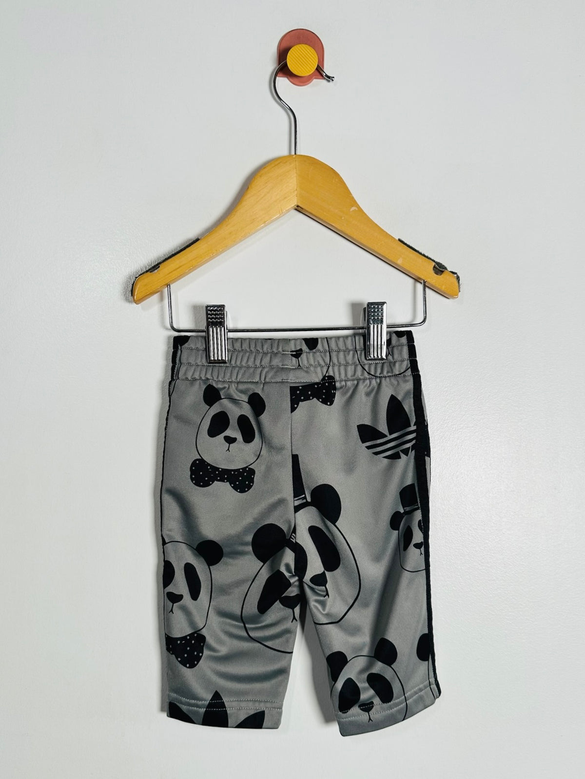 Gray children's shorts with a black panda print and Adidas trefoil logo, hanging on a wooden hanger against a white wall.