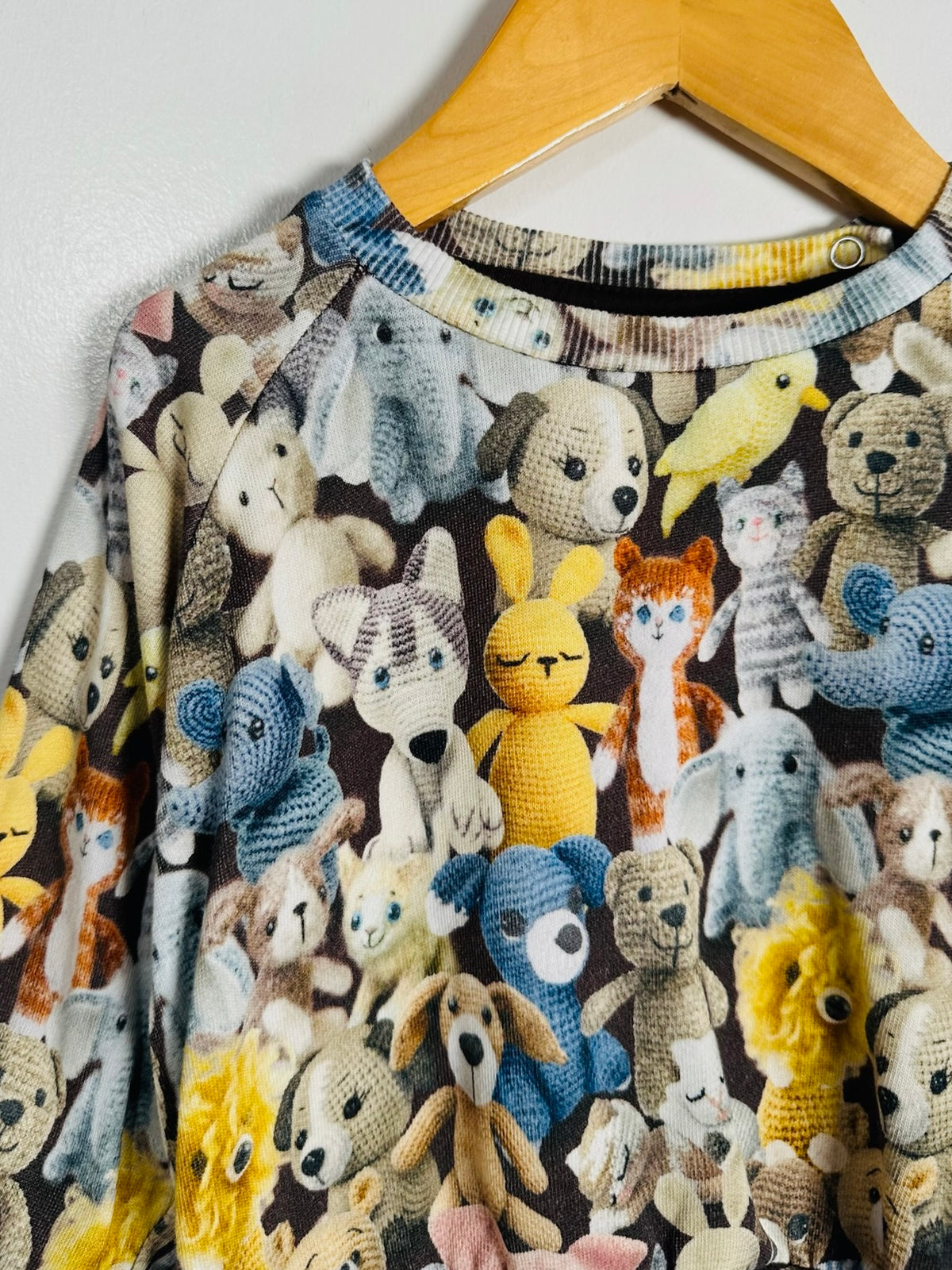 A close-up of a long-sleeve top hanging on a wooden hanger, featuring an all-over print of crocheted and knitted animal toys in yellow, blue, white, and brown on a dark background.