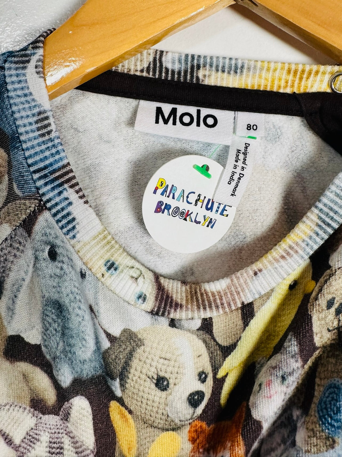 Close-up of a Molo brand sweatshirt hanging on a wooden hanger, featuring a knitted teddy bear pattern and a round 'Parachute Brooklyn' tag inside the collar.