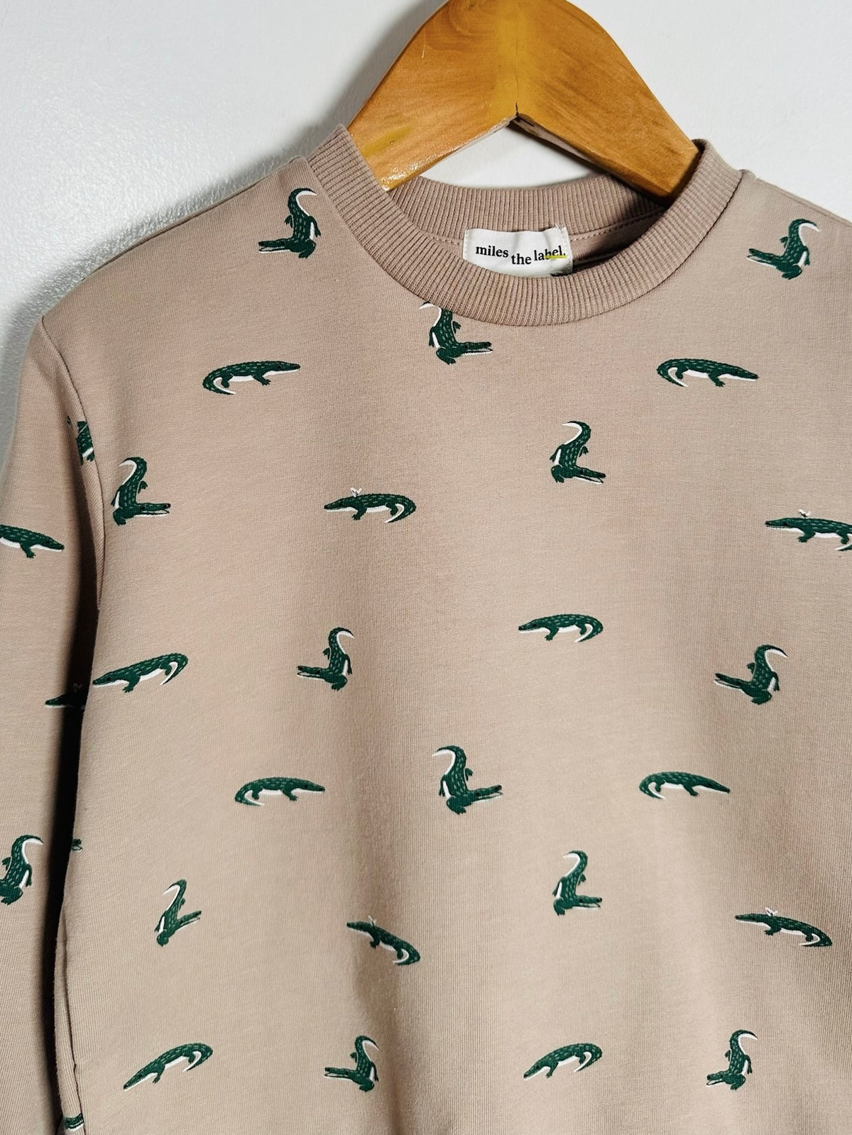 A beige crewneck sweatshirt with an all-over dark green crocodile print, hanging on a wooden hanger against a white wall. The brand label 'milk the rabbit' is visible inside the collar.