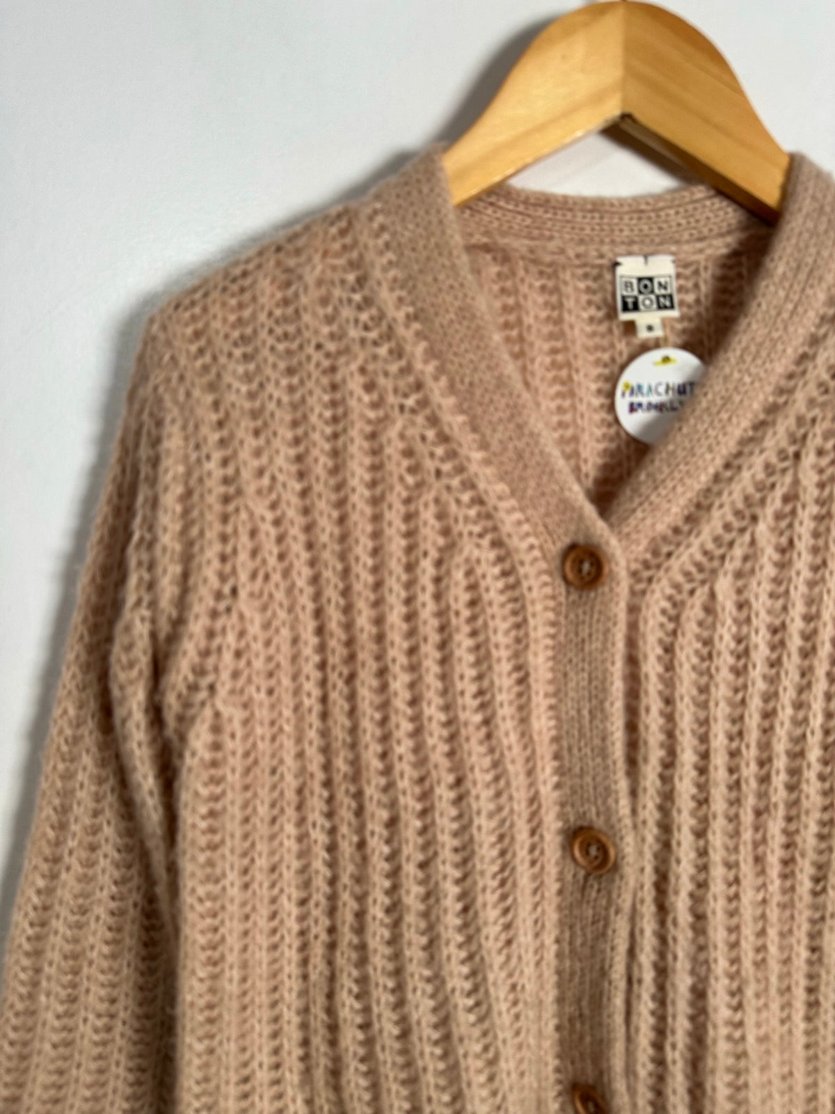 A close-up of a chunky knit cardigan sweater, hanging on a wooden hanger against a plain light background. The cardigan features a V-neck, brown buttons, and a visible 'Macht's Handmade' brand tag.