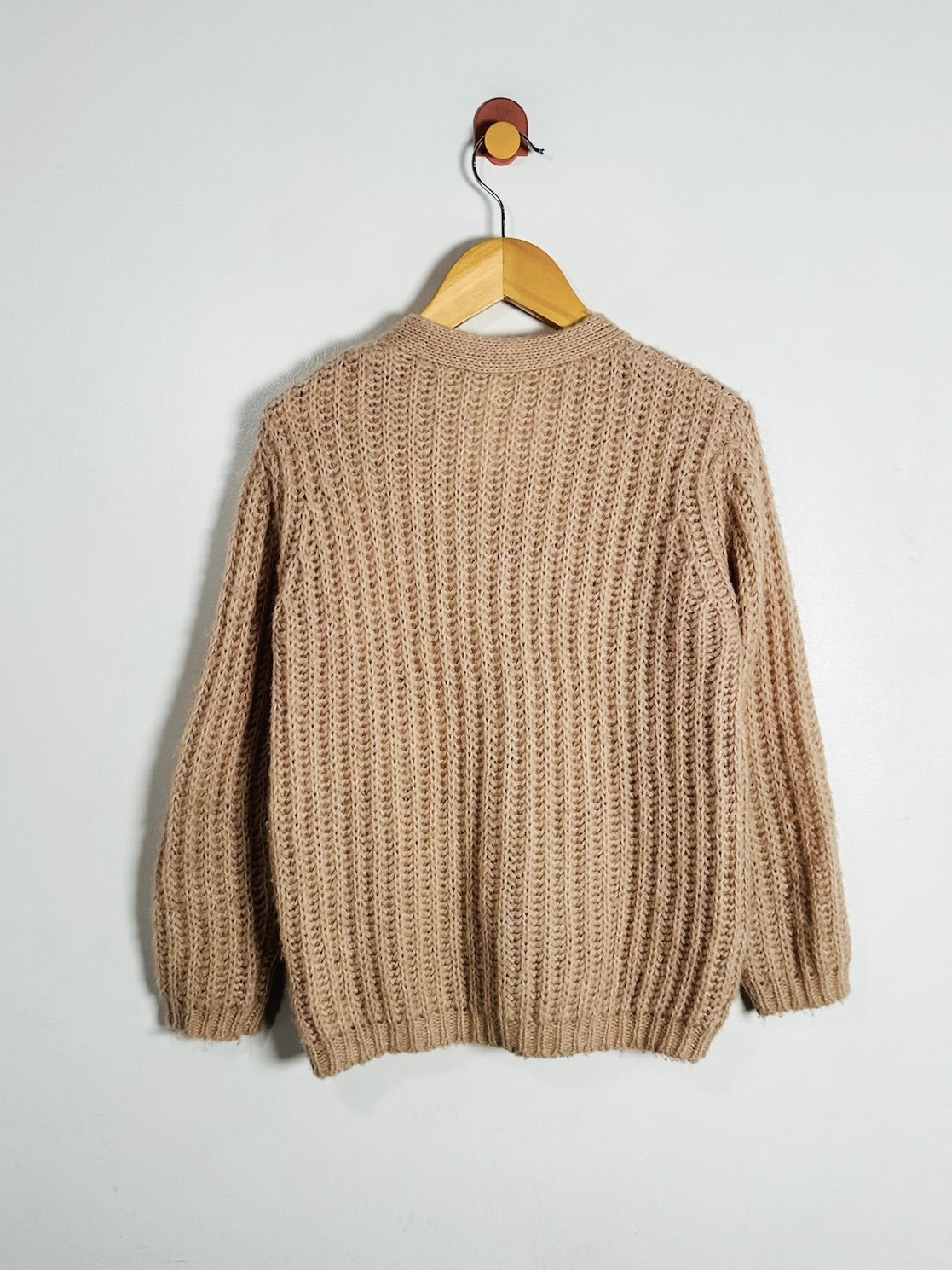 A pink chunky cable-knit sweater hanging on a wooden hanger against a plain white wall.