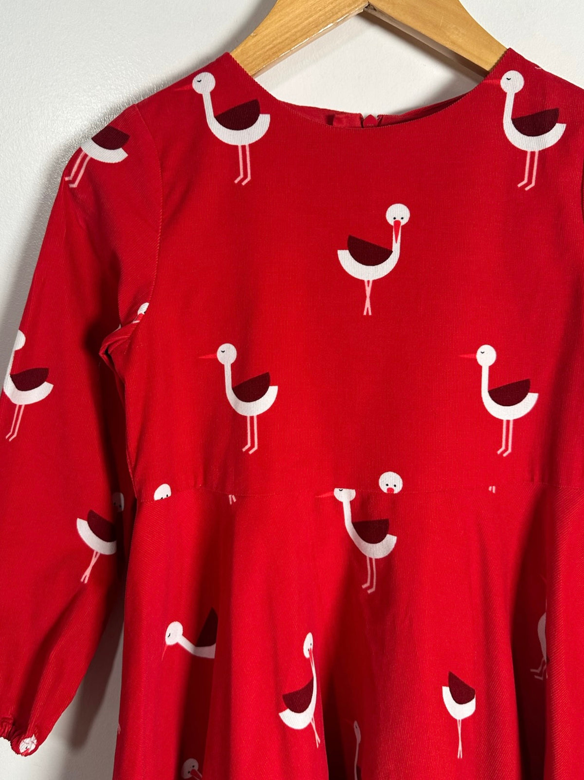 A red long-sleeve dress with a repeating pattern of white and dark red birds, hanging on a wooden hanger against a light wall.