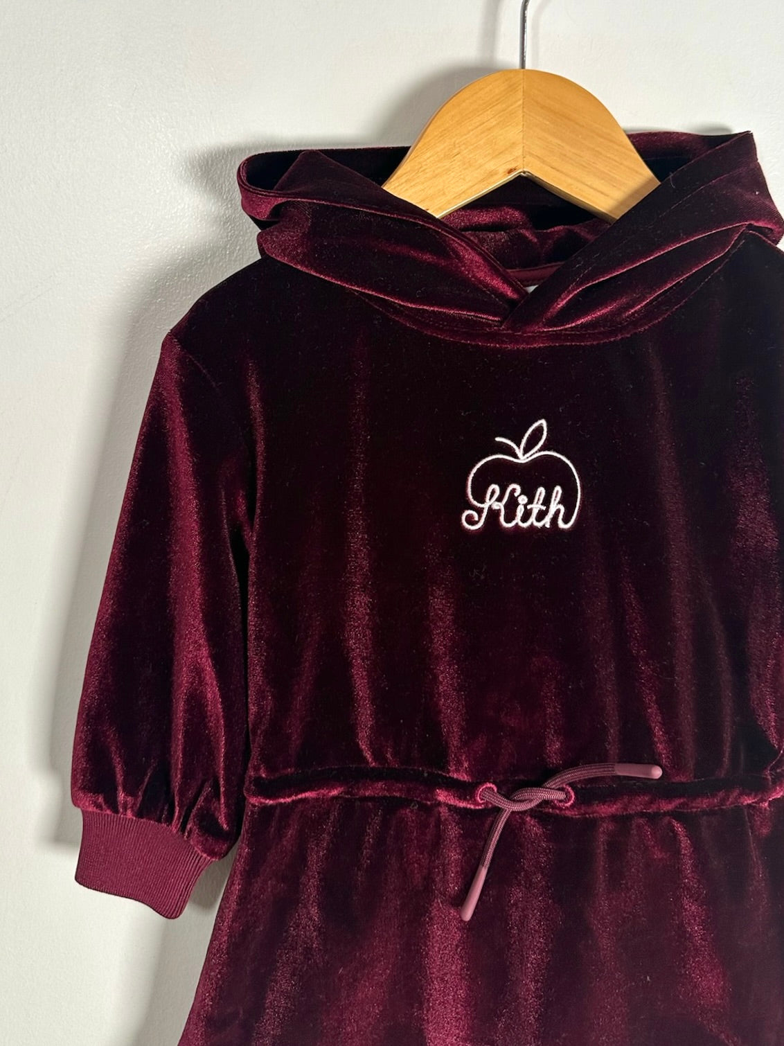 A burgundy velvet hooded dress with a drawstring waist and white embroidered monogram 'Kith' on the chest, hanging on a wooden hanger against a light gray wall.