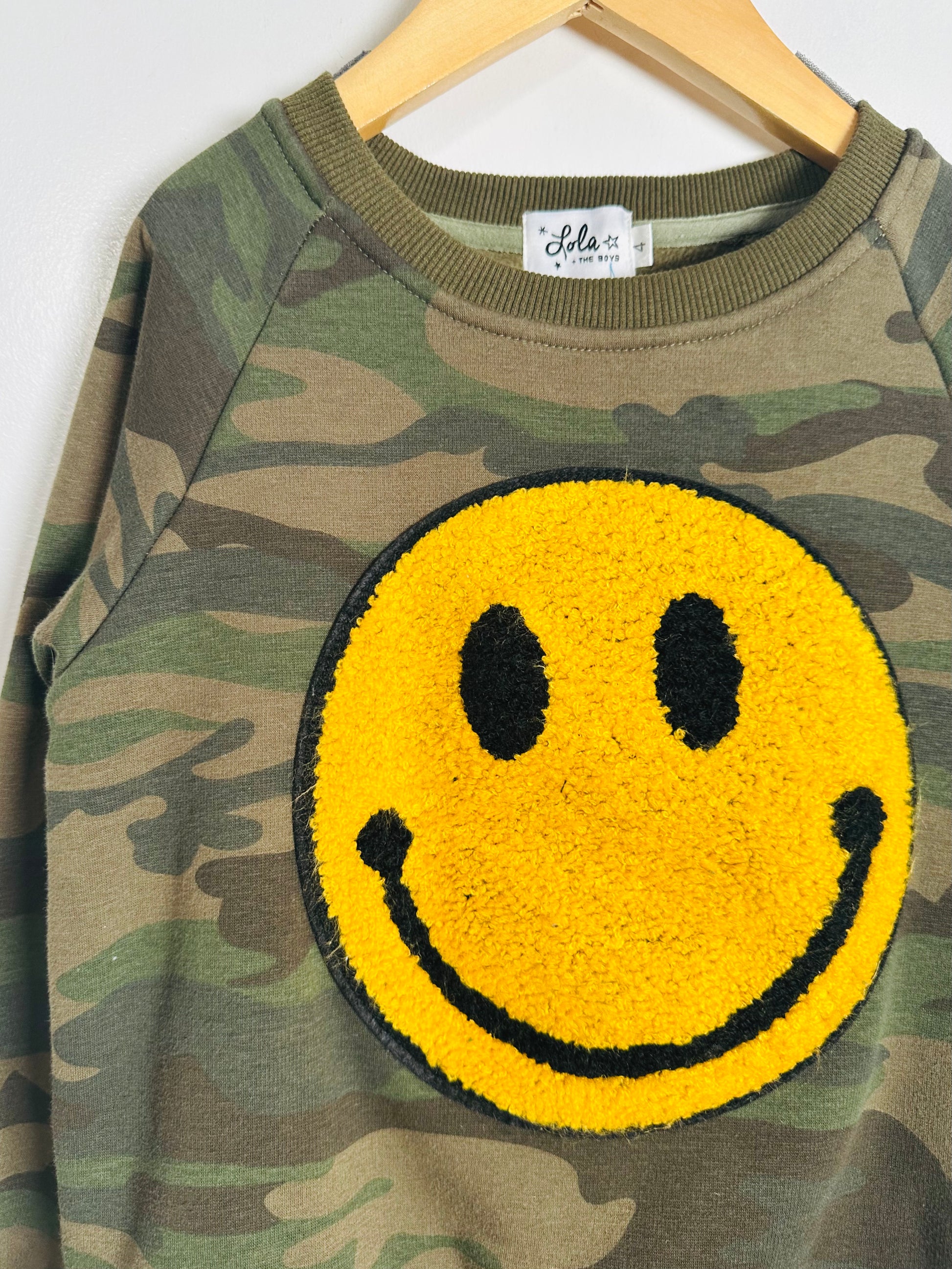 Camouflage sweatshirt with a yellow smiley face patch on a hanger.