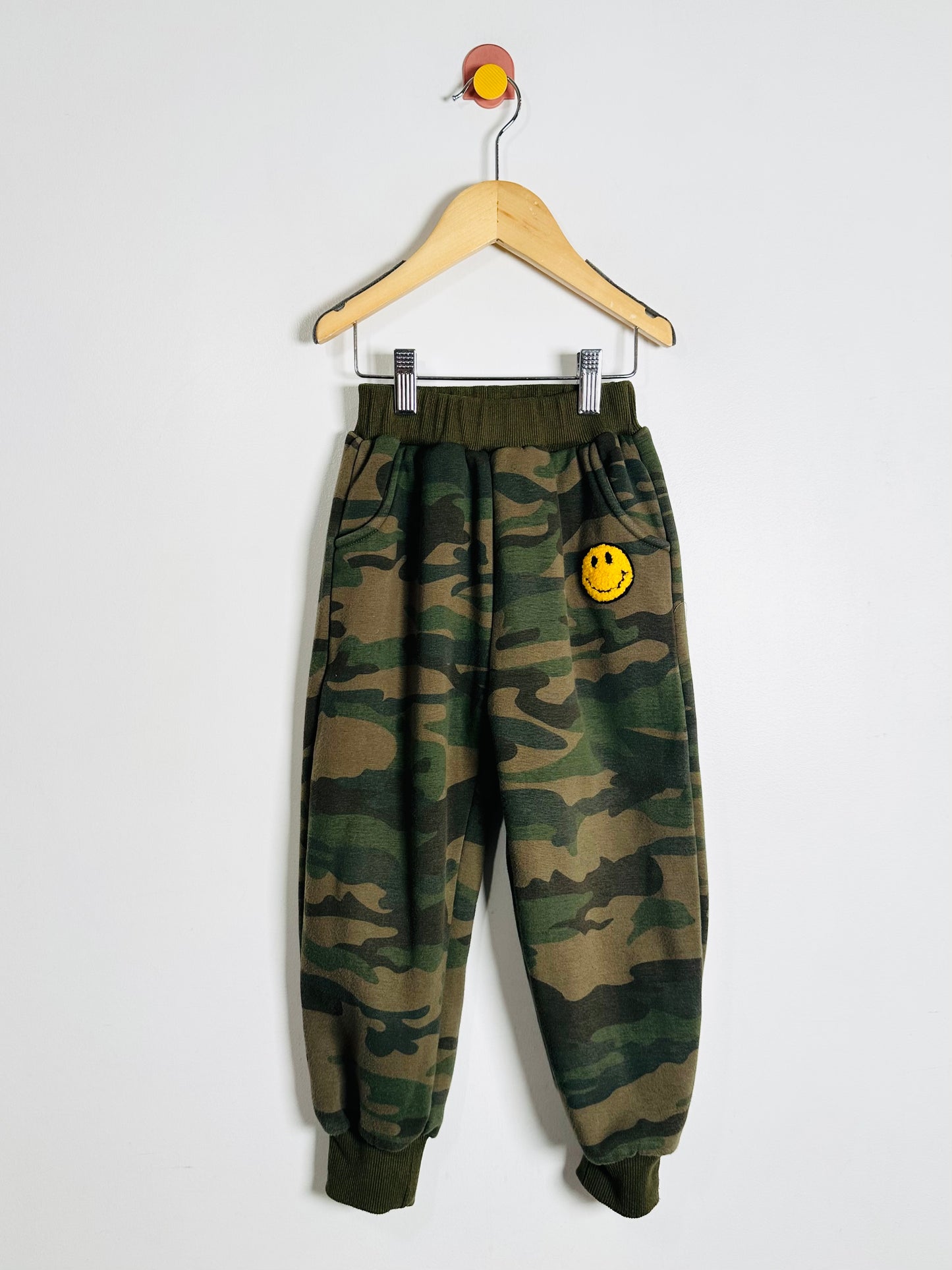 Camouflage pants with a smiley face patch on a hanger against a white background