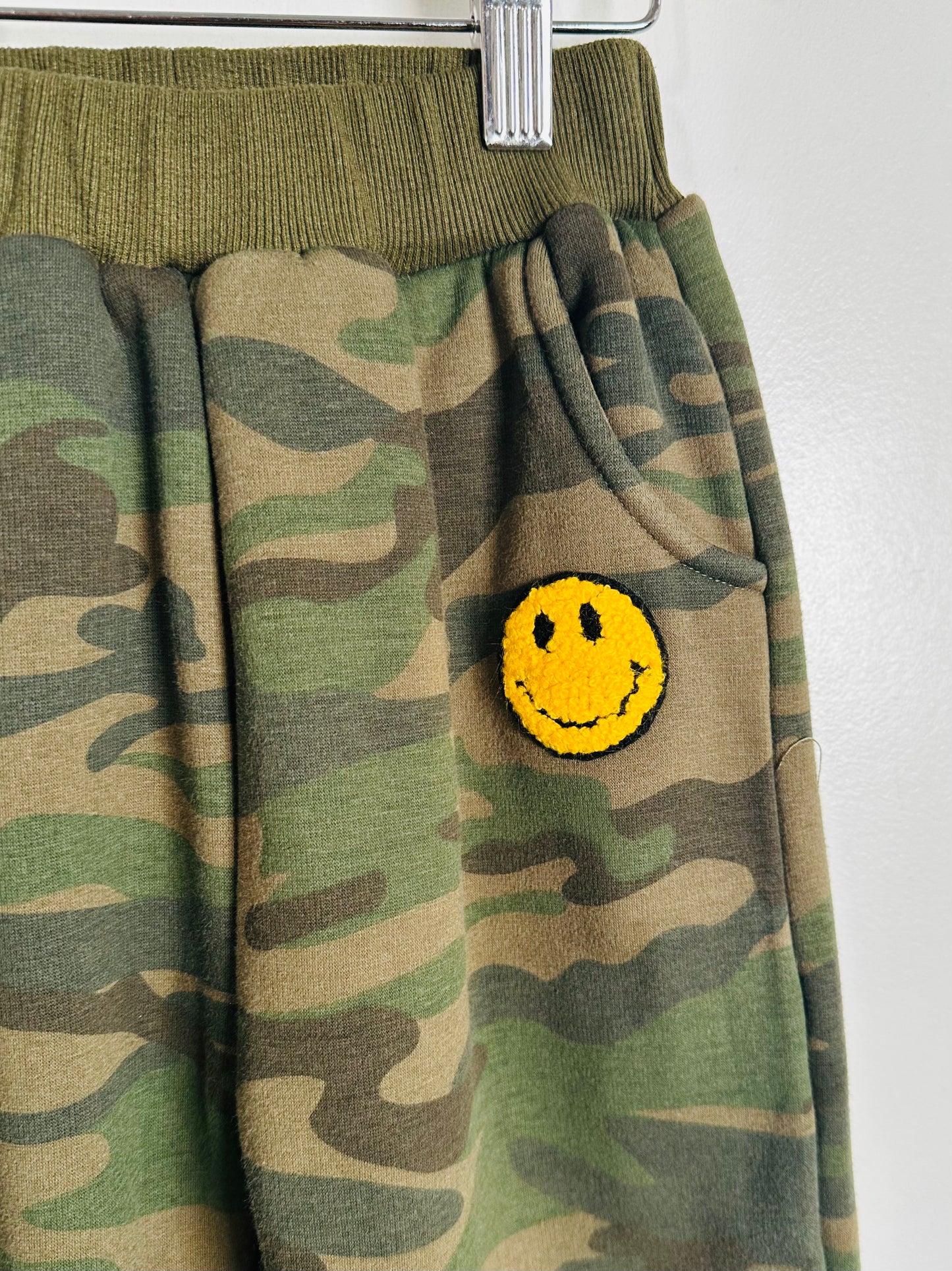 Camouflage pants with a yellow smiley face patch on a white background