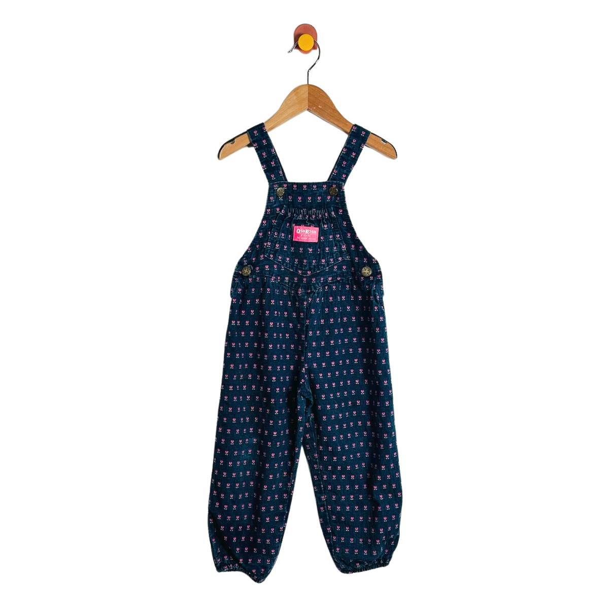 A pair of toddler's dark blue denim overall jumpsuit with a small white and yellow pattern, hanging on a wooden hanger against a white background.