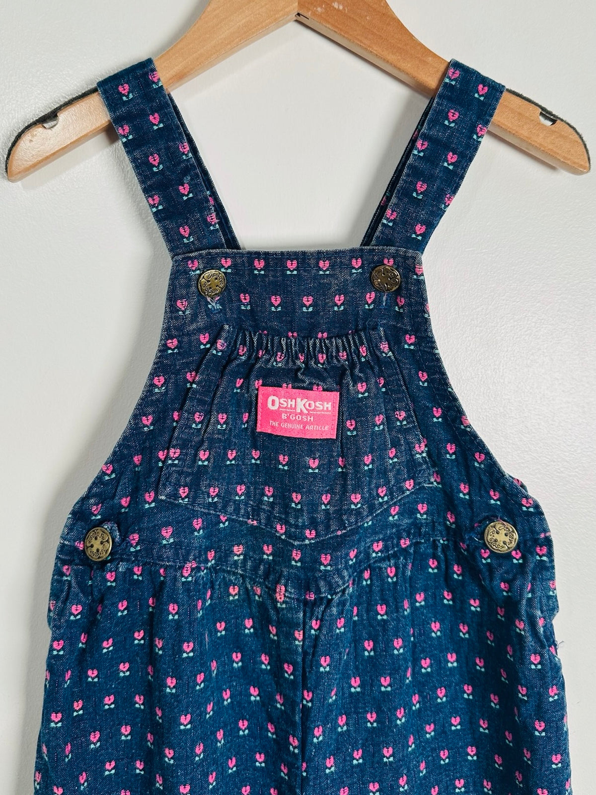 A pair of dark blue denim baby overalls with a pink and white floral pattern, hanging on a wooden hanger against a plain light gray wall. The brand label reads 'OshKosh'.
