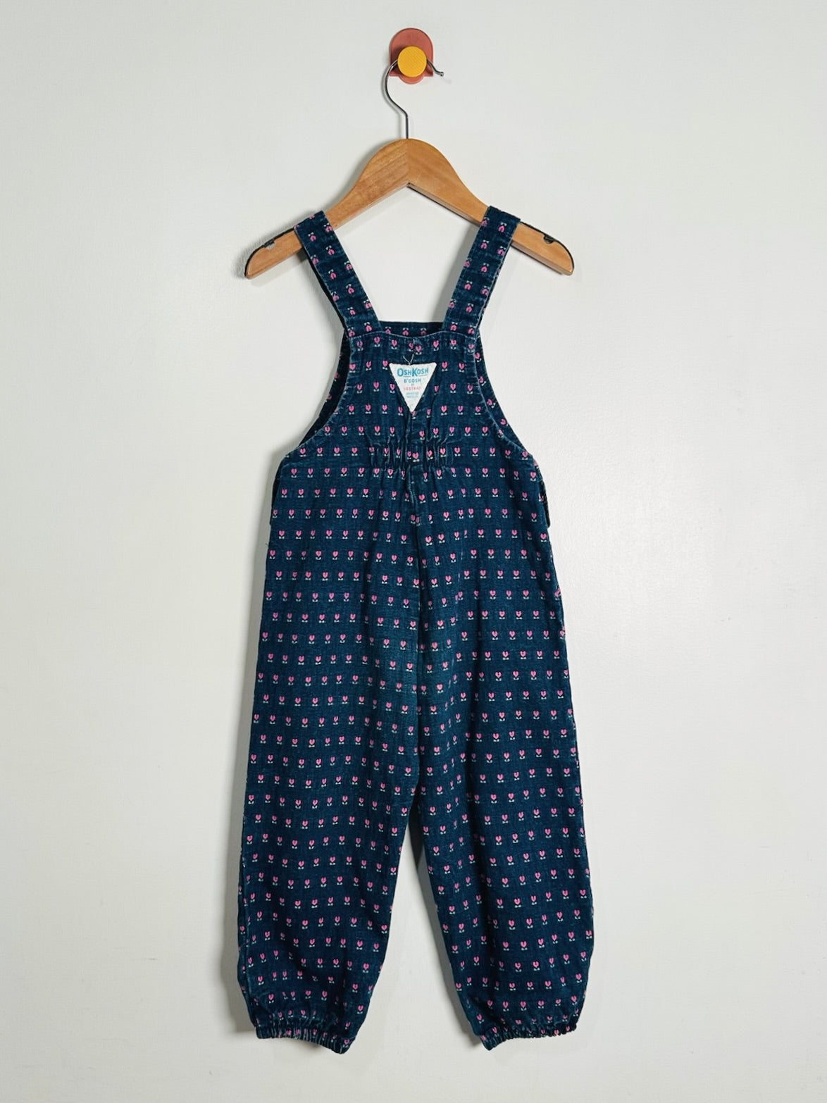 A pair of navy blue children's overall jumpsuit with a small repeating white and pink pattern, hanging on a wooden hanger against a plain off-white wall.