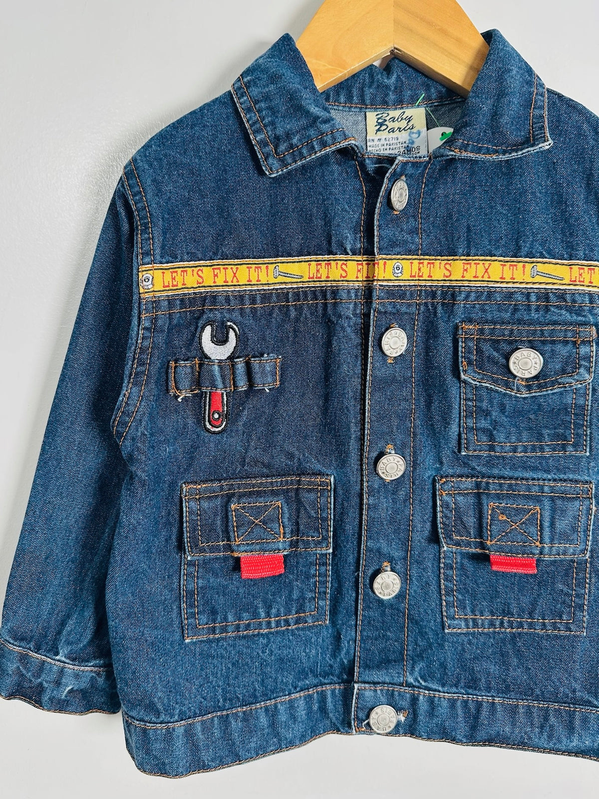 A blue denim jacket for kids, featuring a yellow horizontal stripe across the chest with text, a tool pocket on the left chest with a wrench patch, and red tab accents on the lower pockets, hanging on a wooden hanger against a light gray wall.