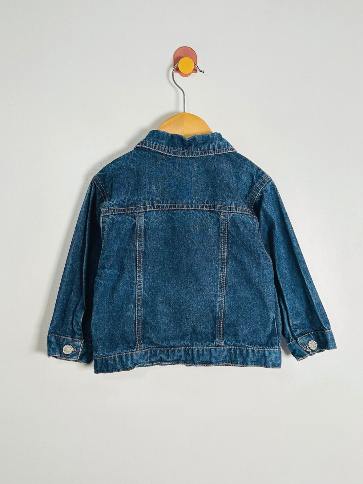 A blue denim jacket shown from the back, hanging on a wooden hanger against a plain light gray wall.