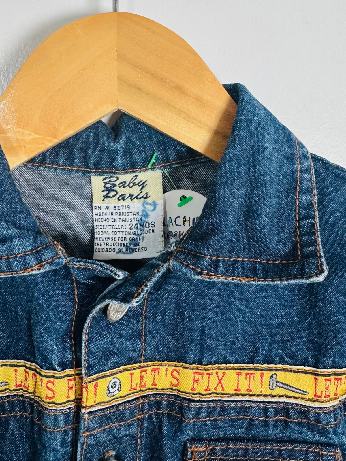 Close-up of a blue denim jacket hanging on a wooden hanger, showing the collar, front placket, and a yellow horizontal stripe with red text reading 'LET'S FIX IT!'. The inner neck label reads Baby Paris.