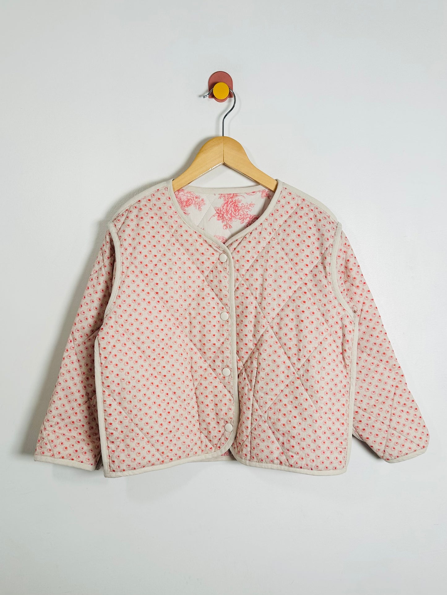 Pink quilted jacket with floral pattern on a hanger against a white background