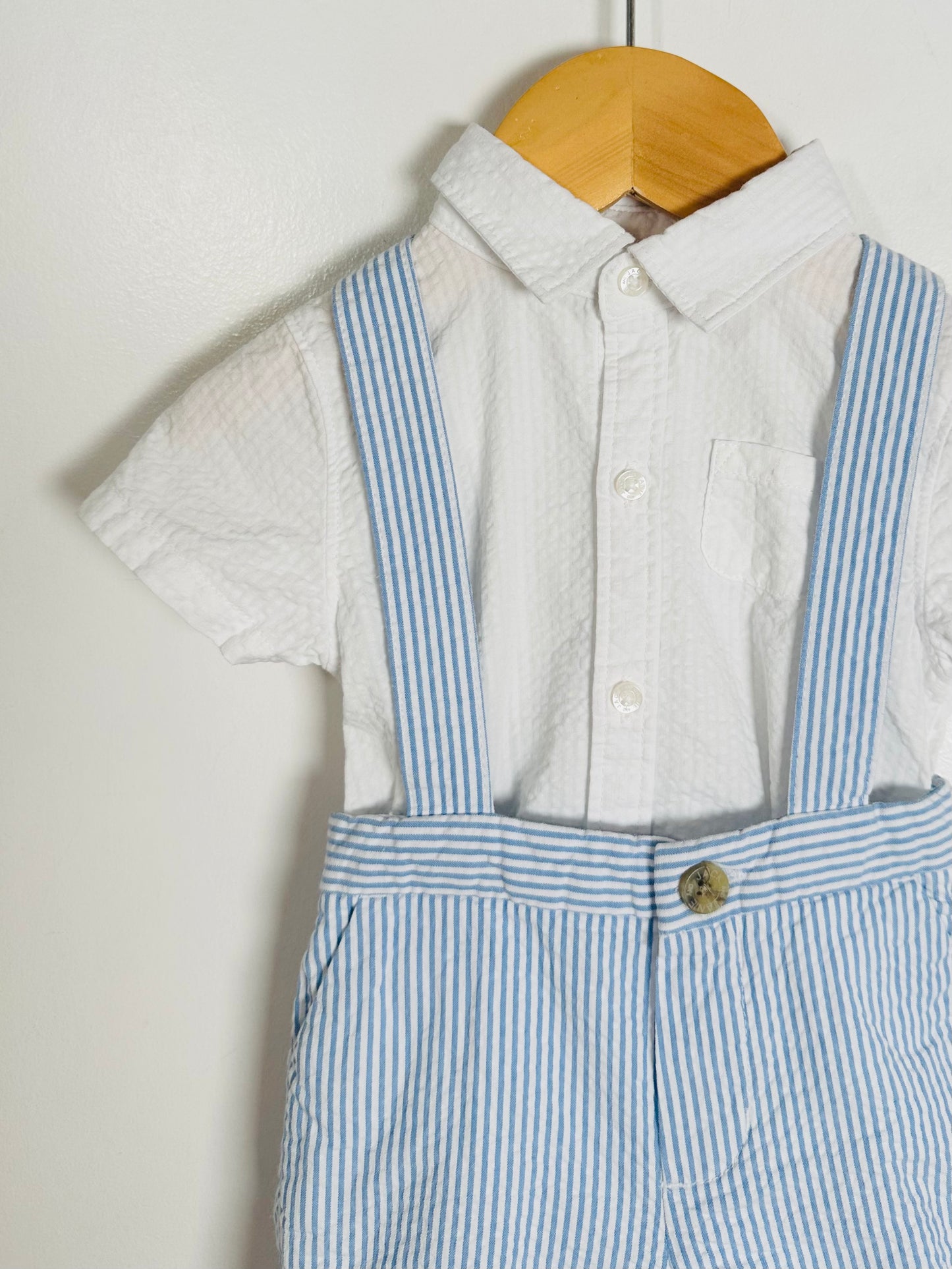 Children's outfit with a white shirt and blue striped overalls hanging on a wooden hanger against a white background.