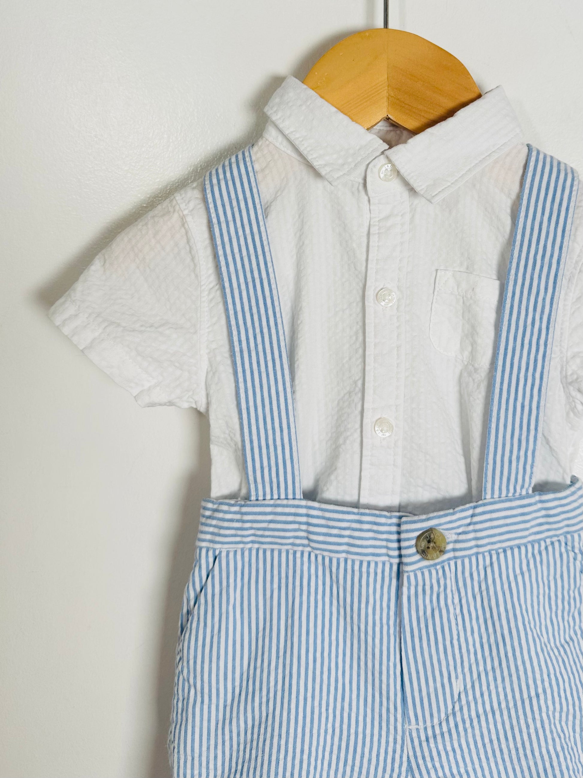 Children's outfit with a white shirt and blue striped overalls hanging on a wooden hanger against a white background.