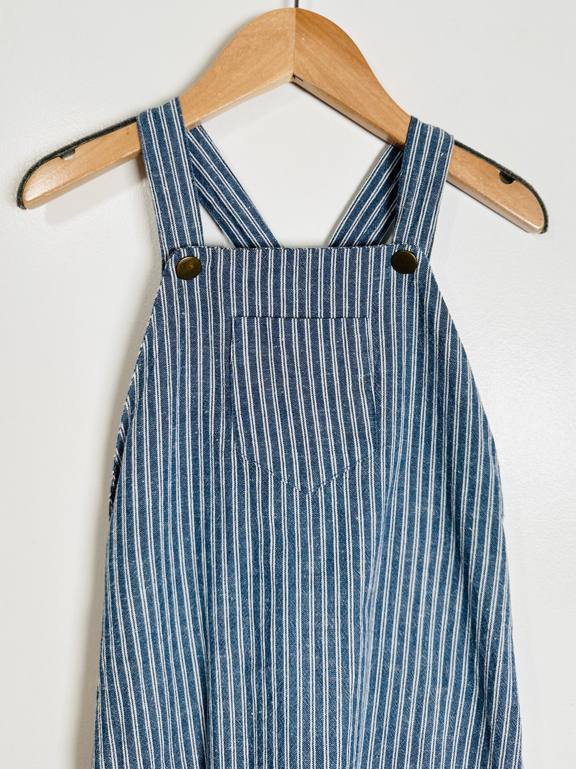 Blue and white striped overalls hanging on a wooden hanger against a white background