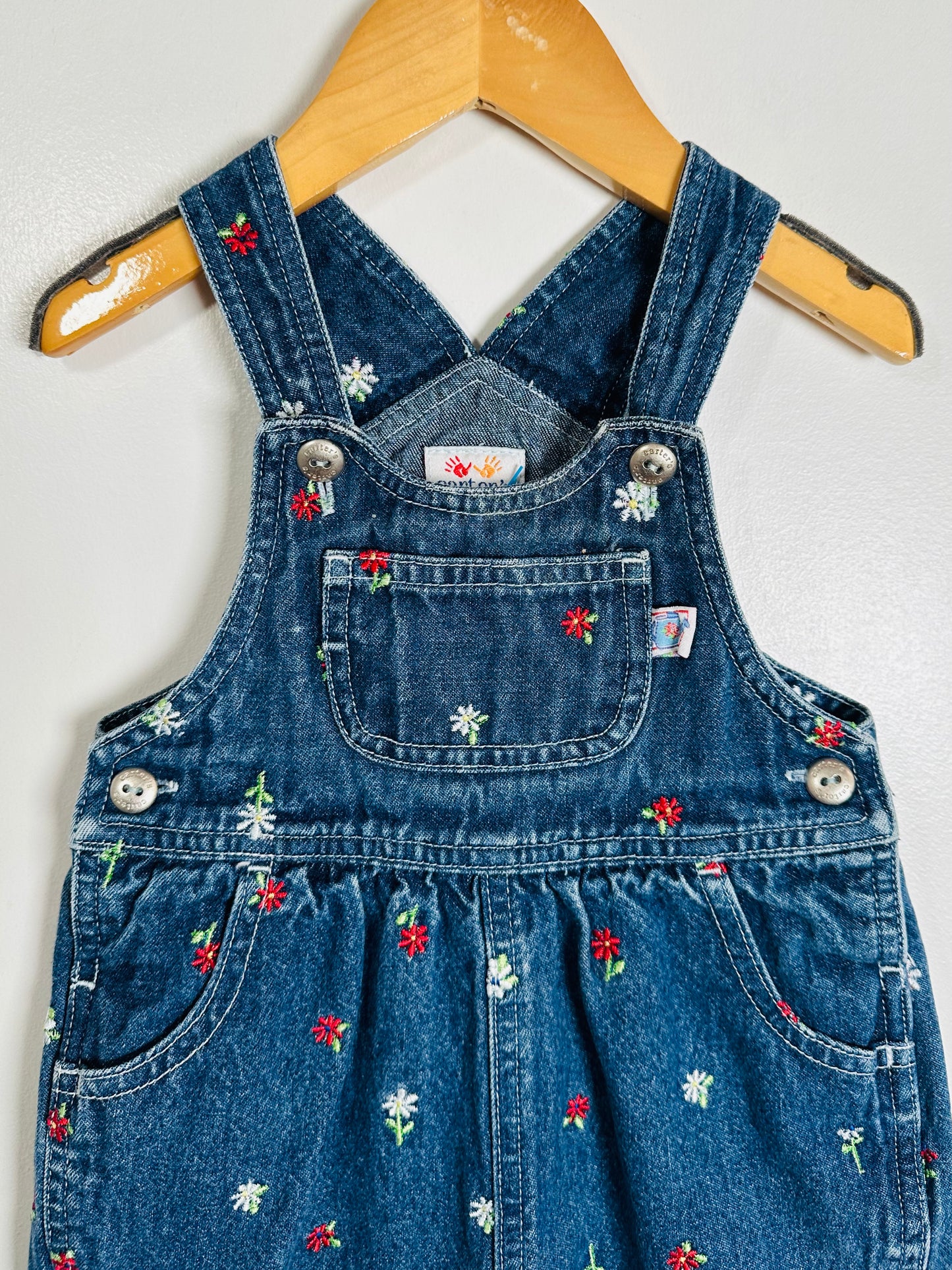 Blue denim overalls with floral embroidery on a wooden hanger against a white background