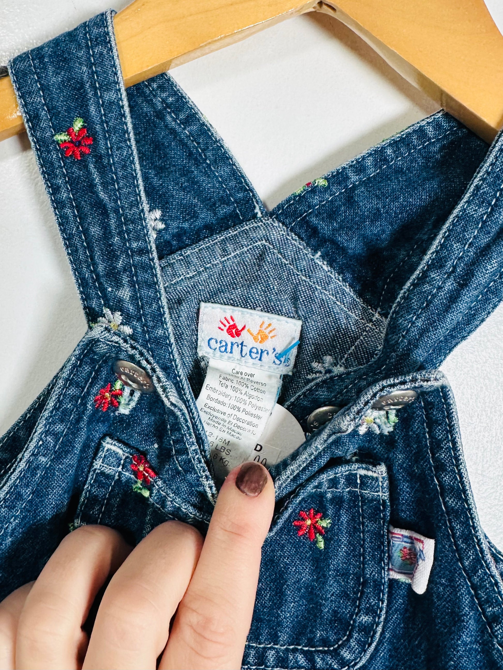 Close-up of a child's denim overalls with floral embroidery and a Carter's tag, held by a hand.