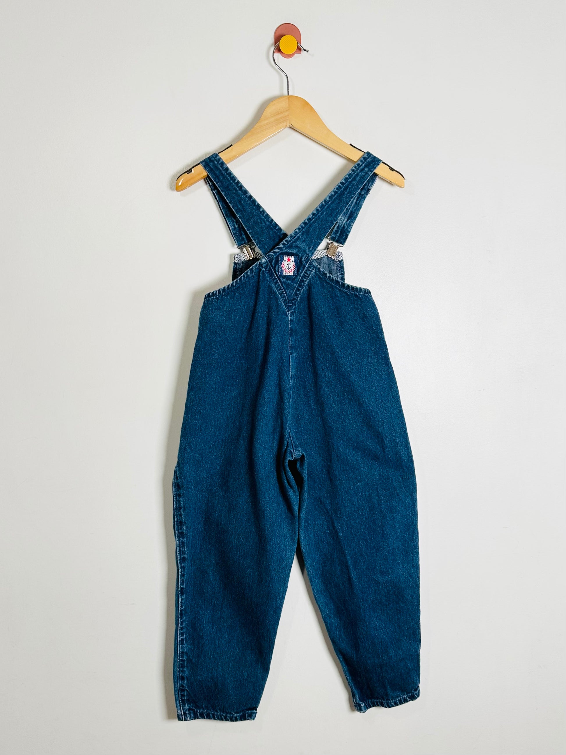 Blue denim overalls hanging on a wooden hanger against a white background