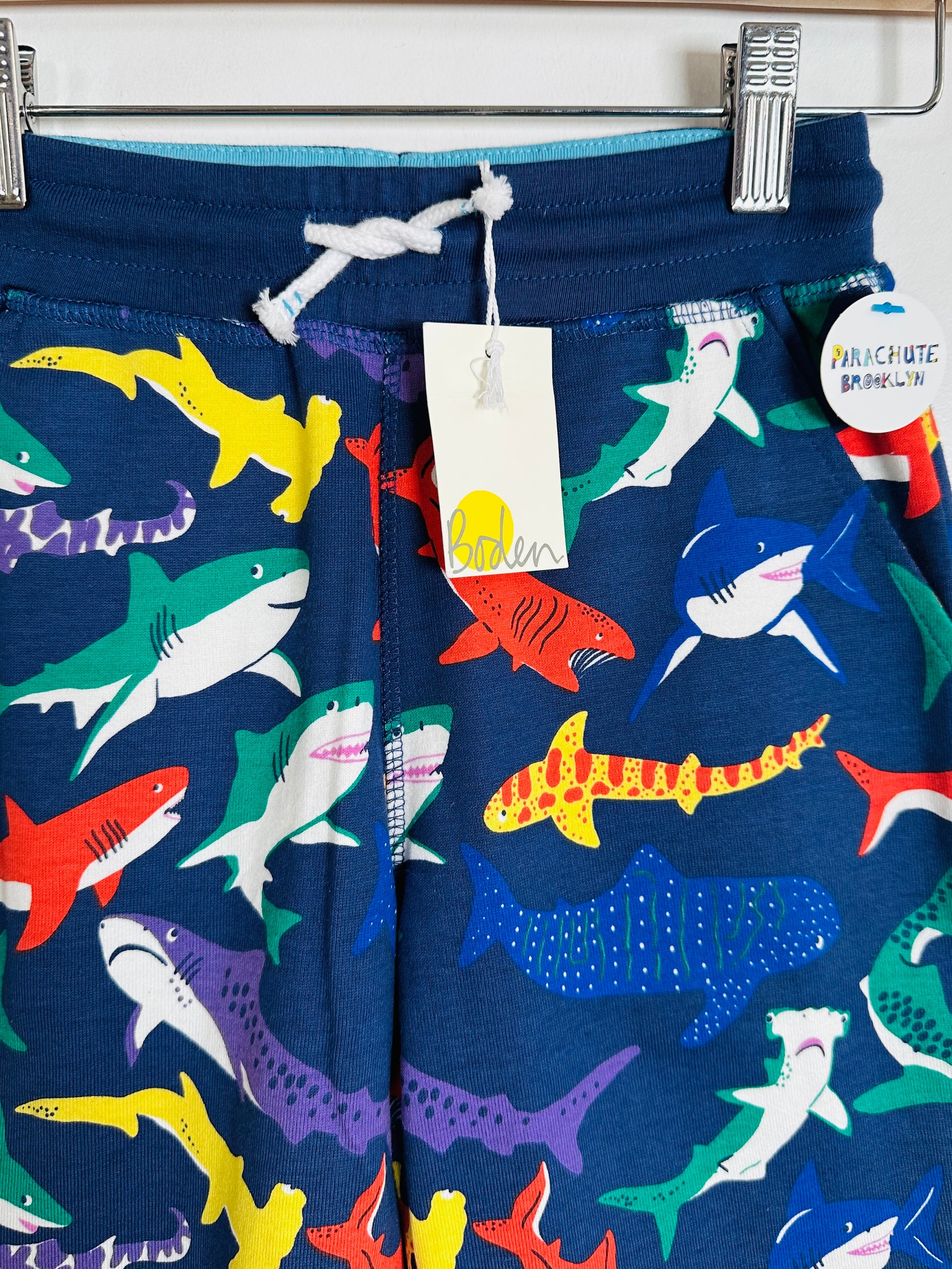Children's shorts with colorful shark pattern on a blue background, hanging on a rack.