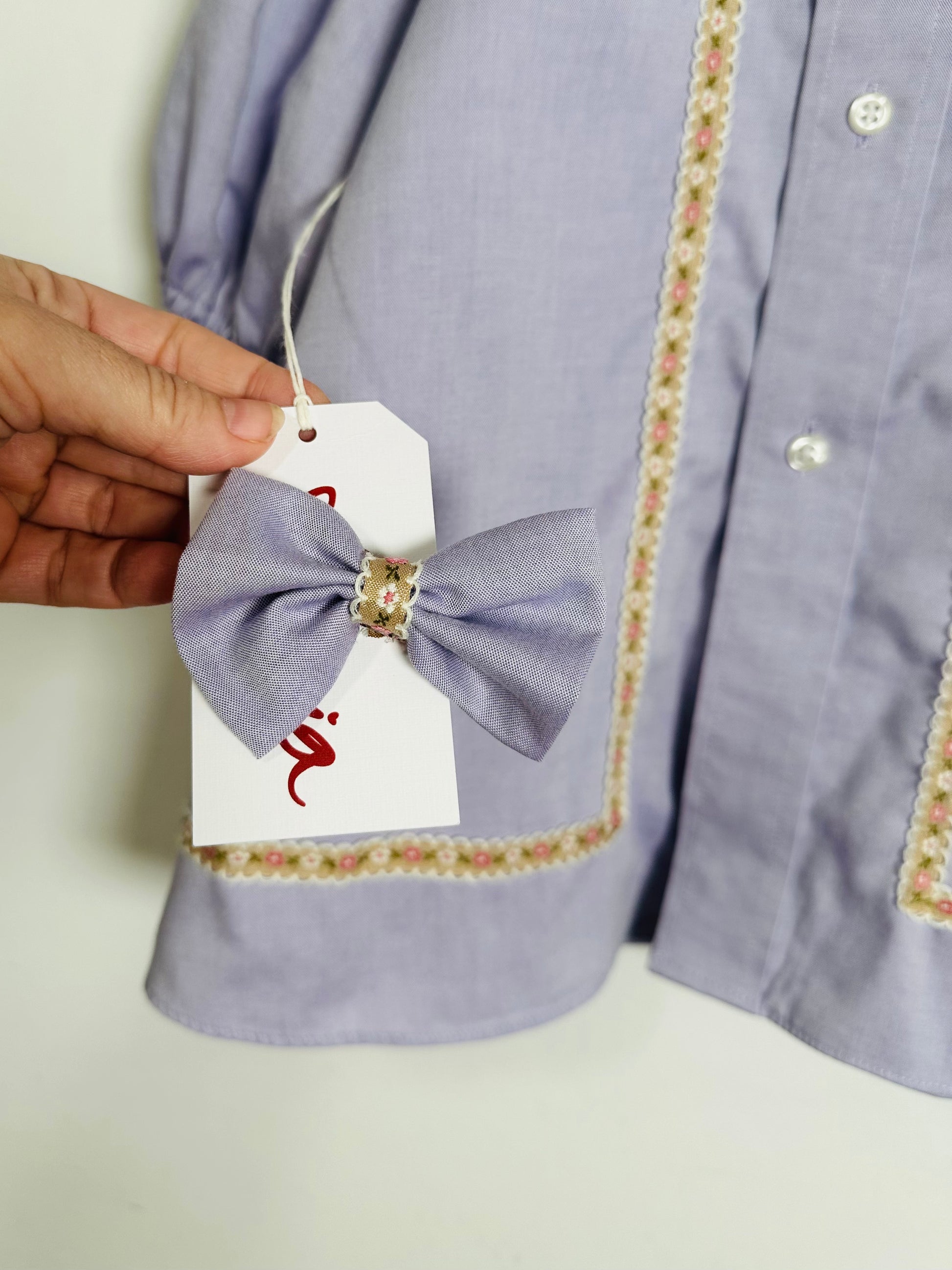 Light purple shirt with floral trim held by a hand, featuring a brand tag with a logo.