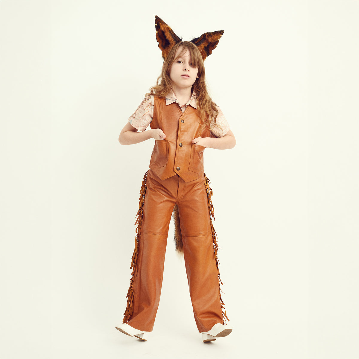 A young girl modeling a brown faux-leather costume set featuring wide-leg pants with fringe, a matching vest, a short-sleeve top, a faux tail, and animal-ear headband against a plain off-white studio background.