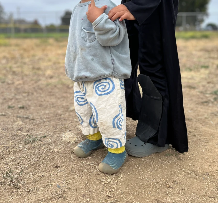A child wearing a light gray sweatshirt, cream pants with blue spiral patterns, and gray slip-on shoes, standing outdoors next to an adult in a black robe.