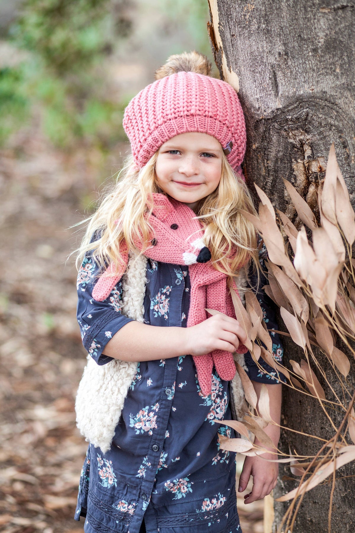 A young girl with blonde hair stands outdoors leaning against a tree trunk, wearing a pink knit beanie with a pom-pom, a matching pink knit scarf with a bunny face detail, and a navy blue floral long-sleeve top with a cream faux-fur vest.