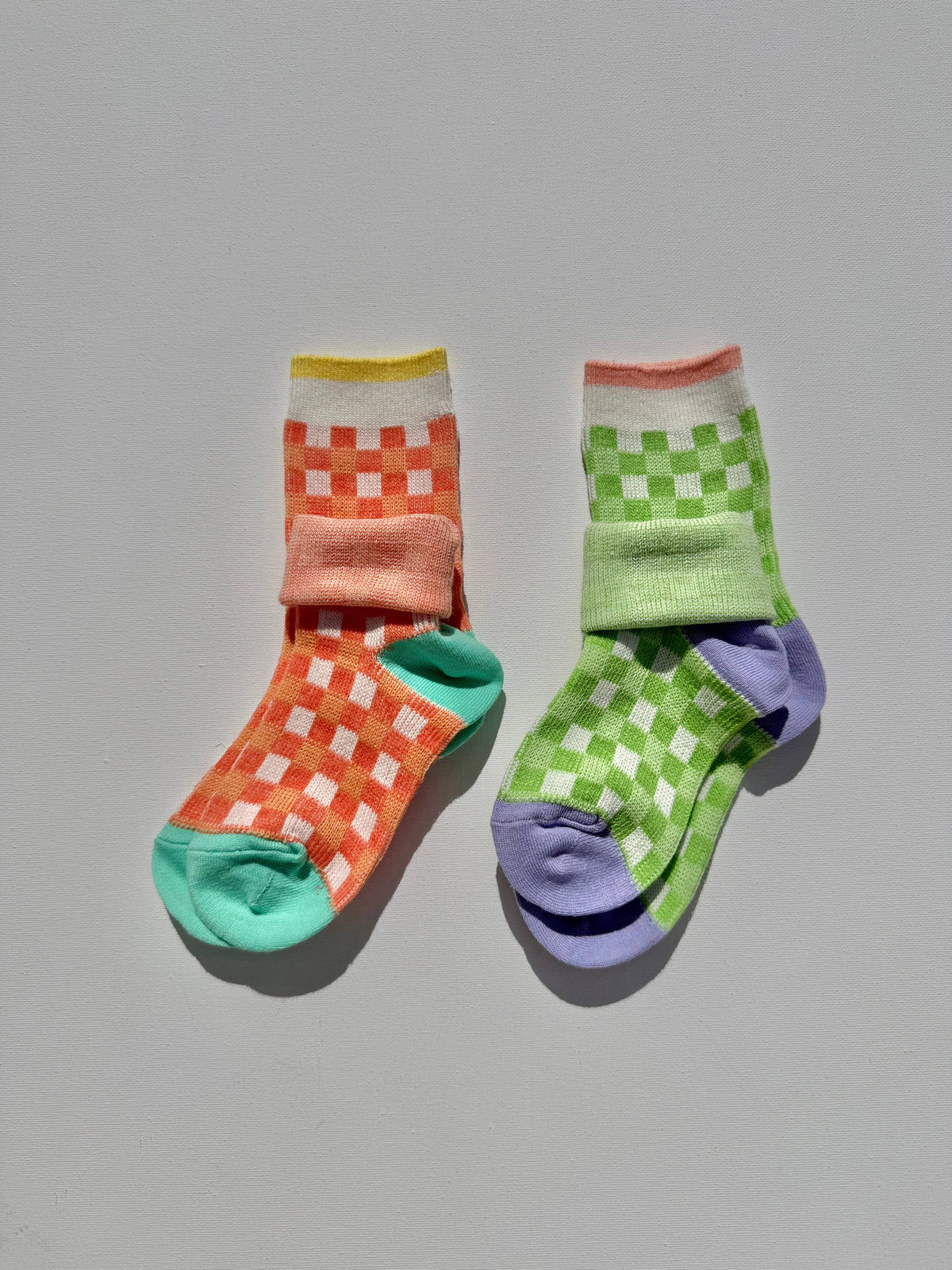 A pair of multicolor patterned crew socks on a neutral gray background. One sock features an orange and white checkered pattern with a mint green toe and heel, while the other features a green and white checkered pattern with a lavender toe and heel.